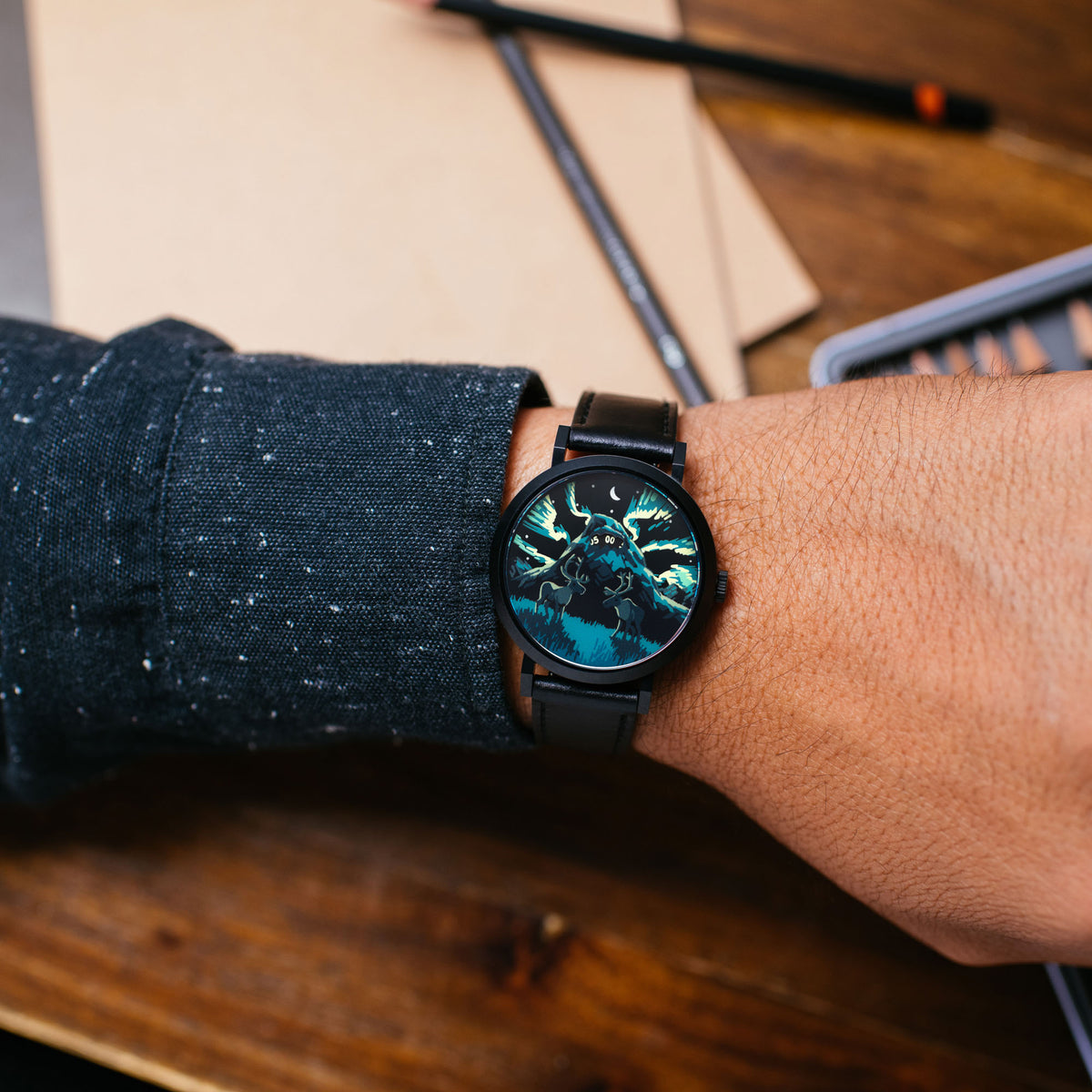 A wristwatch with a black strap and a detailed blue and black dial featuring a wolf design, shown worn on a person's wrist over a dark knit sweater sleeve, set against a wooden desk background with notebooks and a pen.
