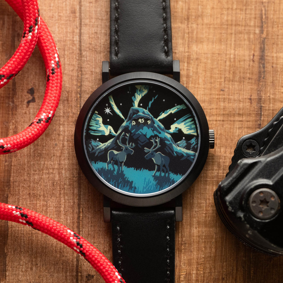 A round analog watch with a black leather strap and a black case, featuring a detailed night-sky illustration with two deer and a mountain on the dial. The watch is placed on a wooden surface next to a red braided cord and a black object.