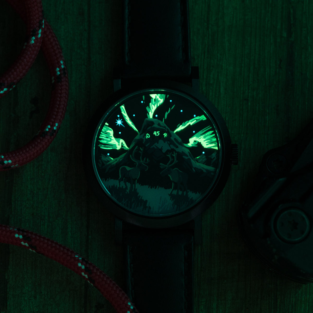 A round watch with a black strap and case, featuring a glowing green night-sky scene with stars and aurora-like light on the dial, photographed on a dark green textured surface with red rope props.