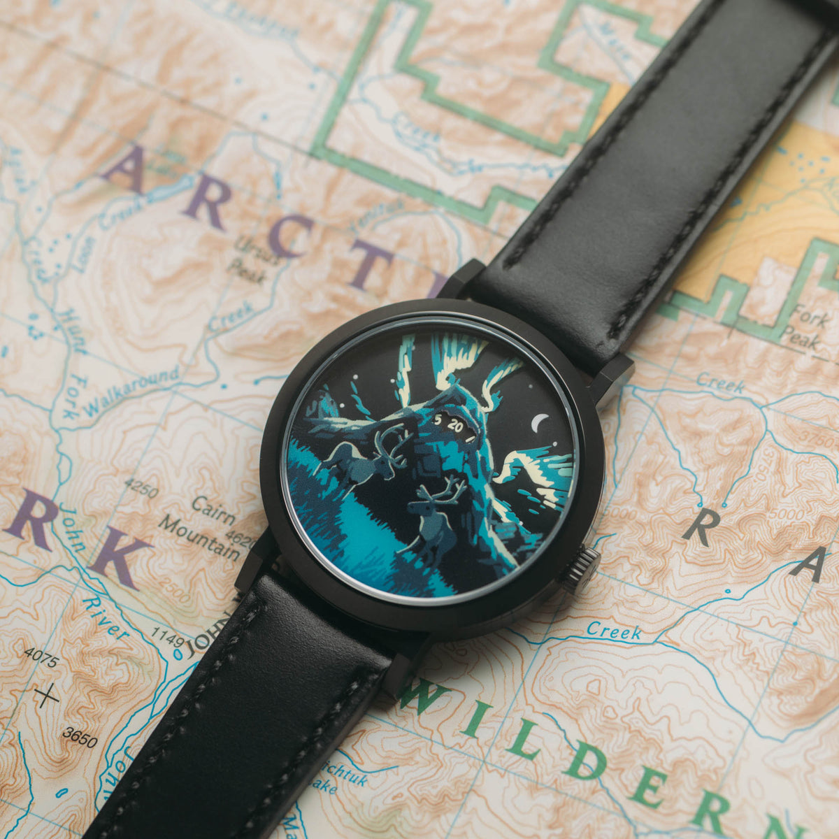 A round analog watch with a black leather strap and a black case, featuring a detailed fantasy illustration of a wolf character on the dial. The watch is placed diagonally on a vintage-style topographic map labeled 'Arctic' and 'Wildern'.
