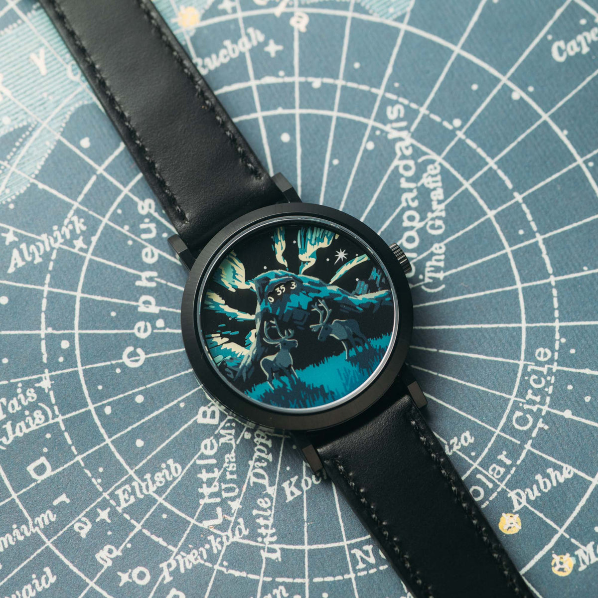 A wristwatch with a black leather strap and a round black case, featuring a detailed blue and black graphic dial of a wolf-like creature. The watch is laid flat on a blue star map background.