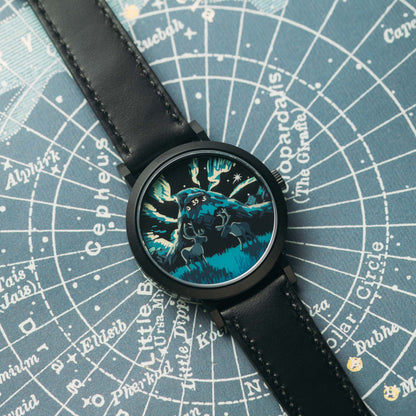 A wristwatch with a black leather strap and a round black case, featuring a detailed blue and black graphic dial of a wolf-like creature. The watch is laid flat on a blue star map background.